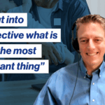 Joe Abeln, smiling in headphones, blue shirt, and a blurred background of a person holding a baby. Quote reads, “It's put into perspective what is really the most important thing.”